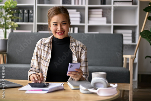 Happy Asian woman managing household expenses and paying bills via mobile banking.