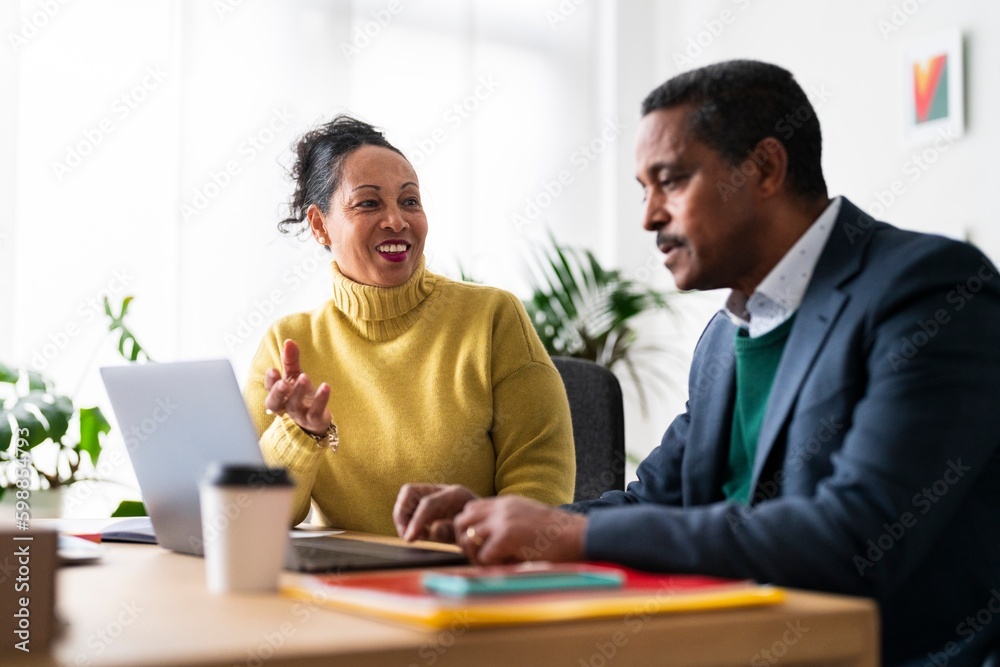 Positive Ethiopian coworkers in smart casual clothes sitting at table near laptop and discussing project details during workday in office Positive Ethiopian coworkers in smart casual clothes sitting at table near laptop and discussing project details during workday in office