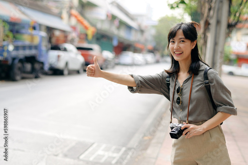 Smiling young Asian woman traveler hitchhiking on a road in the city. Life is a journey concept. © Jirawatfoto
