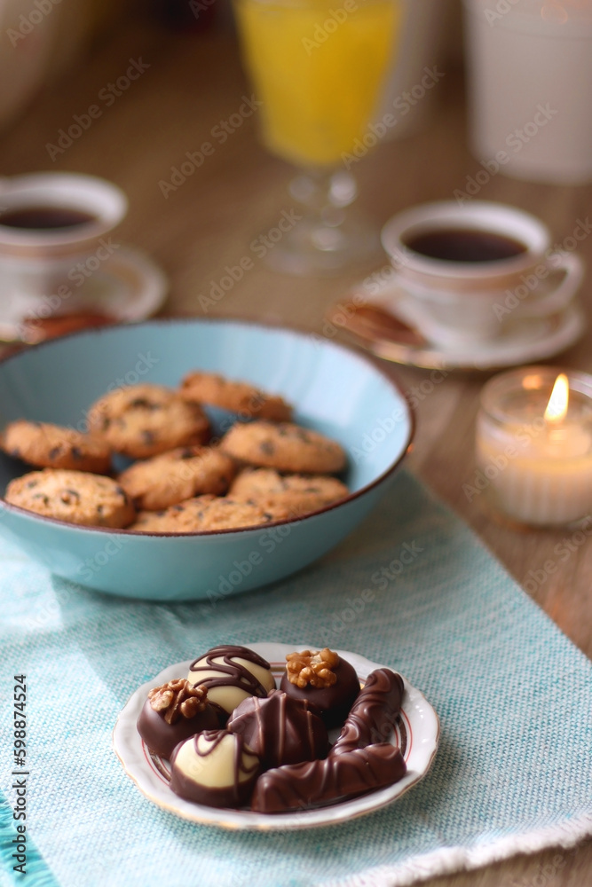 Plate of chocolate pralines, bowl of cookies, cups of tea, glasses of juice and lit candles on the table. Selective focus. Plate of chocolate pralines, bowl of cookies, cups of tea, glasses of juice and lit candles on the table. Selective focus.
