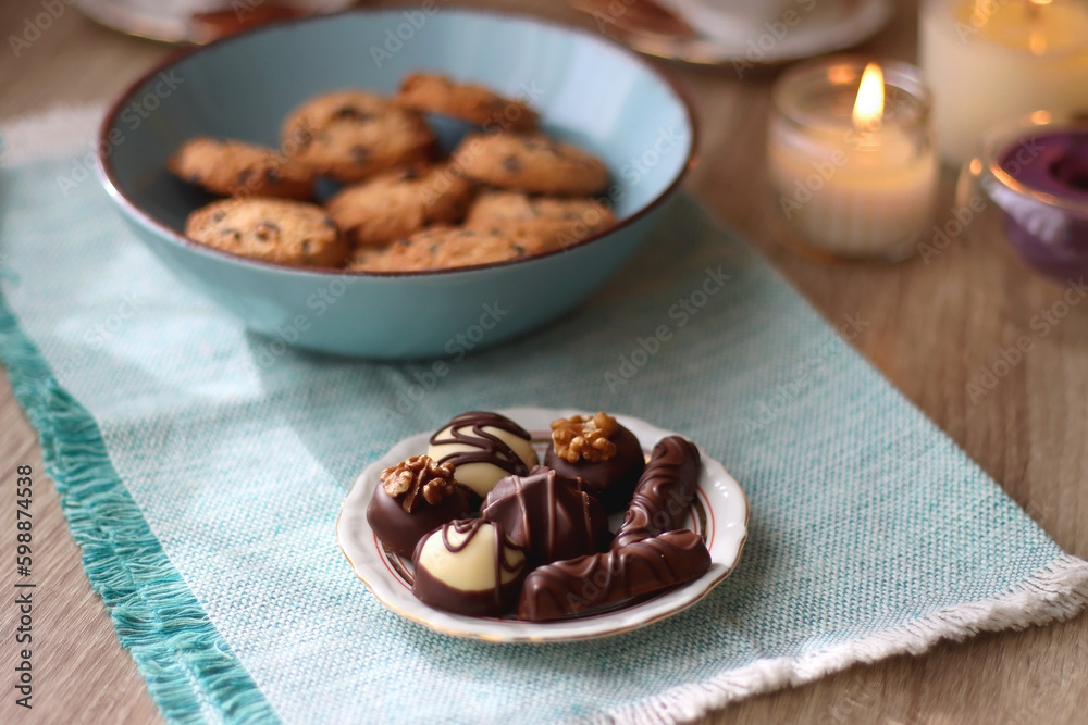 Plate of chocolate pralines, bowl of cookies, cups of tea, glasses of juice and lit candles on the table. Selective focus. Plate of chocolate pralines, bowl of cookies, cups of tea, glasses of juice and lit candles on the table. Selective focus.