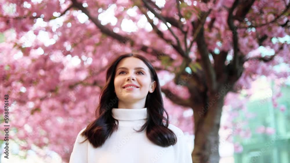 Charming young dark haired woman walking down the city street and enjoying beautiful day with sakura flowers in the background Pretty woman looking at pink blossom at spring morning outdoors Charming young dark haired woman walking down the city street and enjoying beautiful day with sakura flowers in the background Pretty woman looking at pink blossom at spring morning outdoors