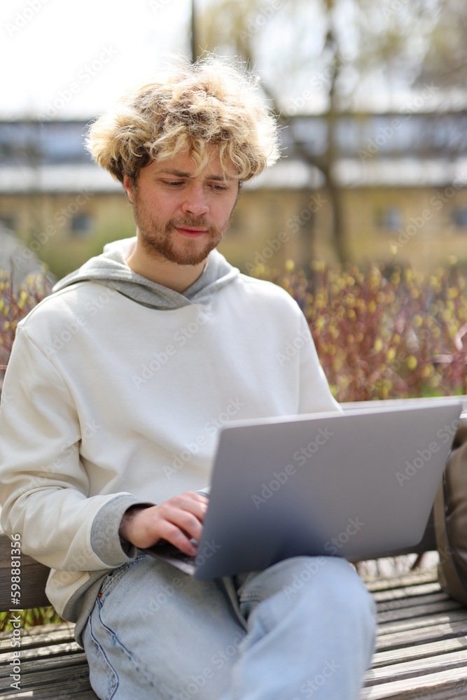 A curly-haired man works on a laptop outside, a student sits on a bench in the park. A curly-haired man works on a laptop outside, a student sits on a bench in the park.