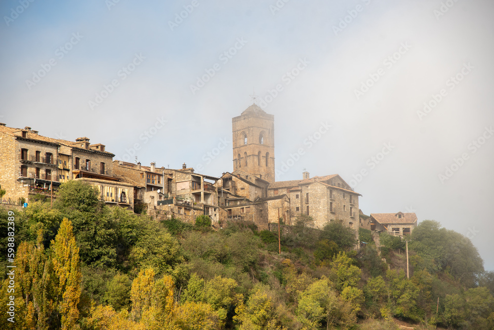 Panoramic view of the touristic village of Ainsa in Huesca surrounded by a natural landscape with its church on top of the hill and small brick and wooden houses during the sunset of a sunny day 