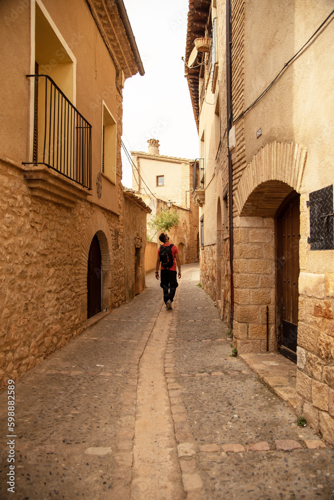 Tourist with a backpack and an orange T-shirt walking through the streets of the tourist village of Alquezar observing its architecture of small brick houses in Huesca, Aragon.
