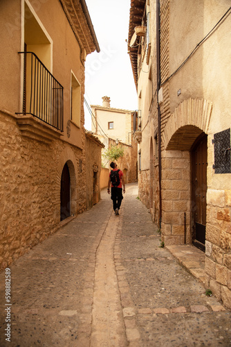 Tourist with a backpack and an orange T-shirt walking through the streets of the tourist village of Alquezar observing its architecture of small brick houses in Huesca, Aragon. © Safi