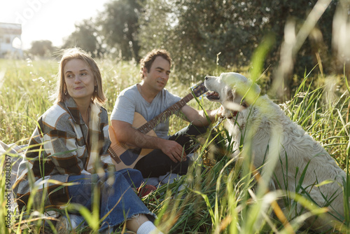 Happy caucasian couple with their labrador dog sitting on the lawn in summer nature with guitar