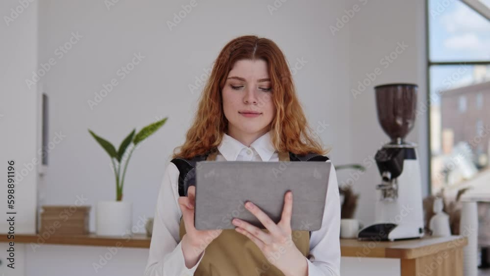 Female successful barista girl, cafe owner is taking online orders on tablet looking at the camera in a cozy, bright cafe. Portrait of charming red-haired girl in restaurant. Entrepreneurs, business. Female successful barista girl, cafe owner is taking online orders on tablet looking at the camera in a cozy, bright cafe. Portrait of charming red-haired girl in restaurant. Entrepreneurs, business.