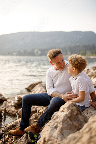 Brunette father and his blondie curly hair son sitting at looking to each other at the stones of Italian Garda lake  © Natalia