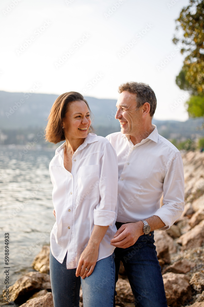 Obraz premium Portrait of stylish casual couple in love in white embracing and looking at each other at the Italian lake stone coast 