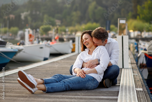 Stylish couple in love in white shirts hugging each other smiling at the wooden pier at Italian Garda lake 