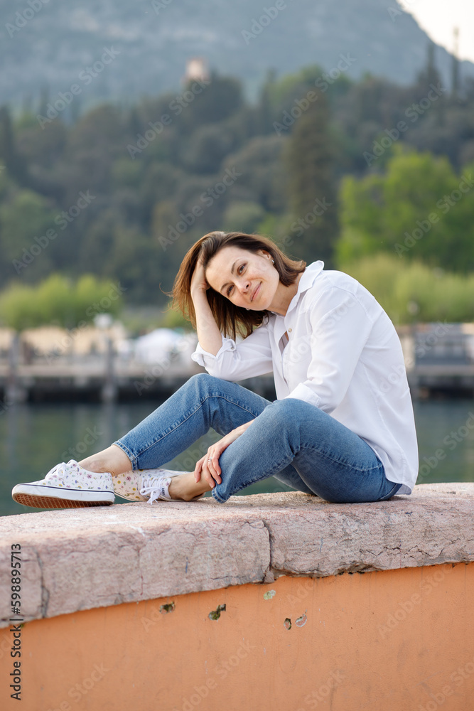 Pretty brunette woman sitting at the railing at the Italian Garda lake port in white shirt enjoying her vacation 