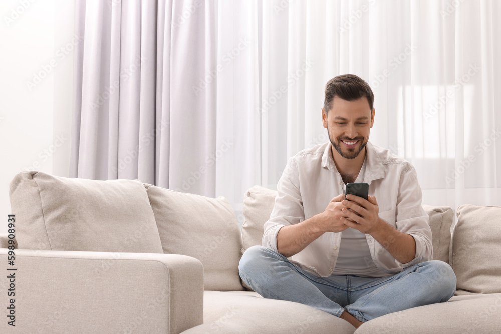 Man using smartphone on sofa near window with beautiful curtains in living room. Space for text