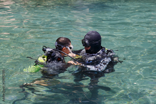 Diving with aqualung lesson, instructor giving scuba diving lessons to a kid. Trainer teaching a little boy to dive. Two scuba divers dressed in diving suit is preparing to dive in shallow sea.