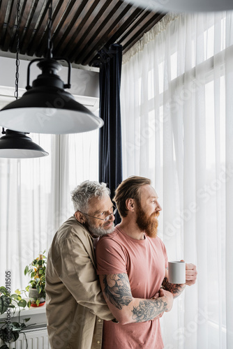 Mature gay man hugging tattooed partner with cup of coffee at home. 