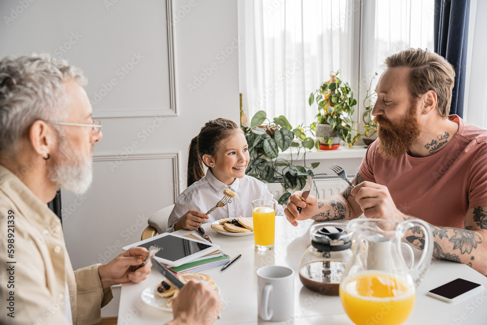 Smiling girl sitting near homosexual parents and tasty breakfast at home. 