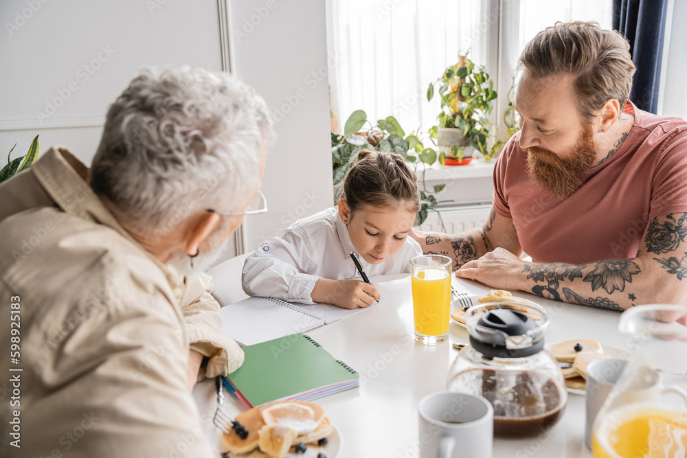 Preteen girl doing homework near gay parents and breakfast at home. Preteen girl doing homework near gay parents and breakfast at home.