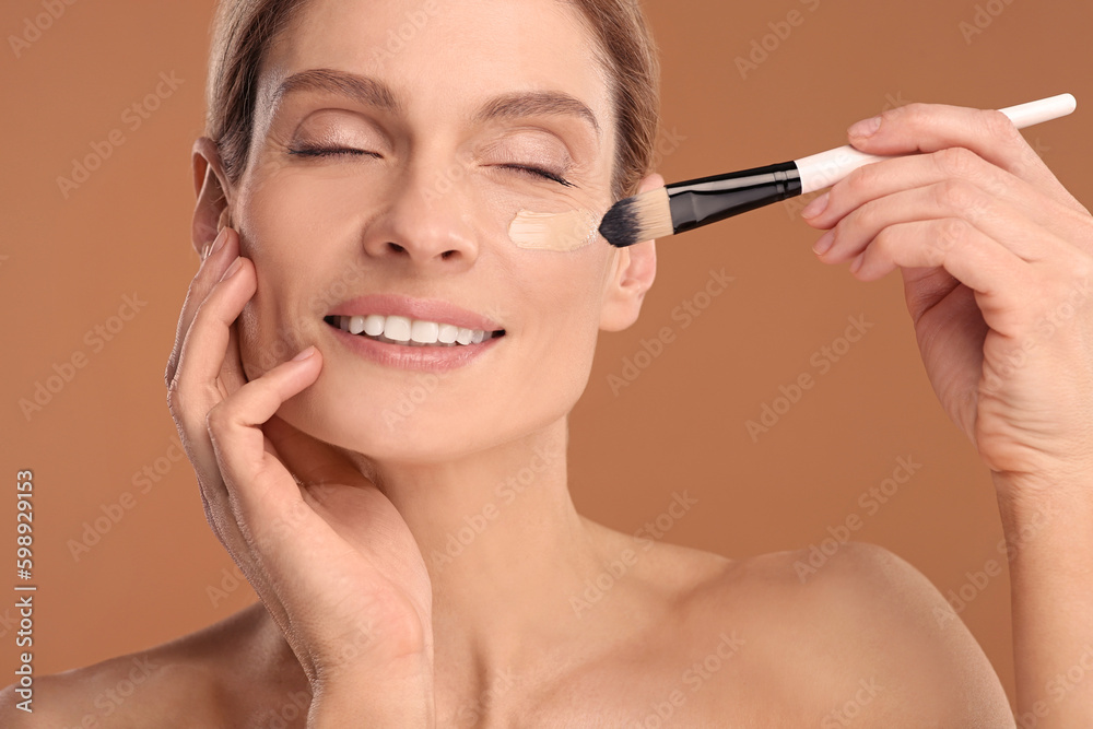 Fototapeta premium Woman applying foundation on face with brush against brown background