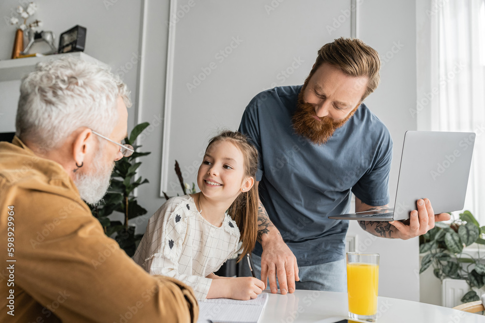 Smiling girl doing homework with same sex parents during online education at home. Smiling girl doing homework with same sex parents during online education at home.