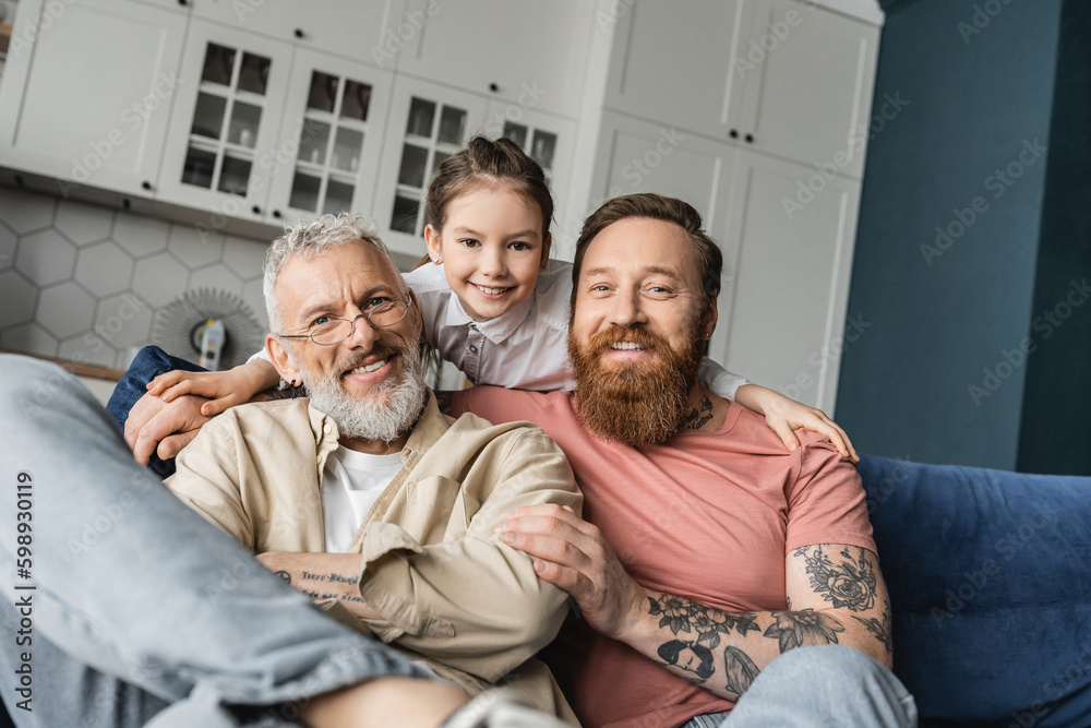 Smiling girl hugging tattooed and happy gay fathers on couch at home. 
