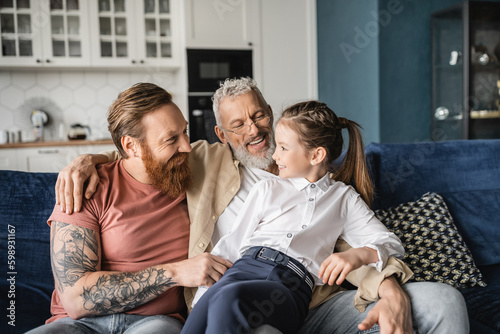 Canvas Print Cheerful gay parents looking at adopted daughter on couch at home