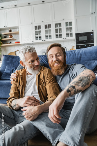 Positive same sex couple sitting near couch on floor at home. 
