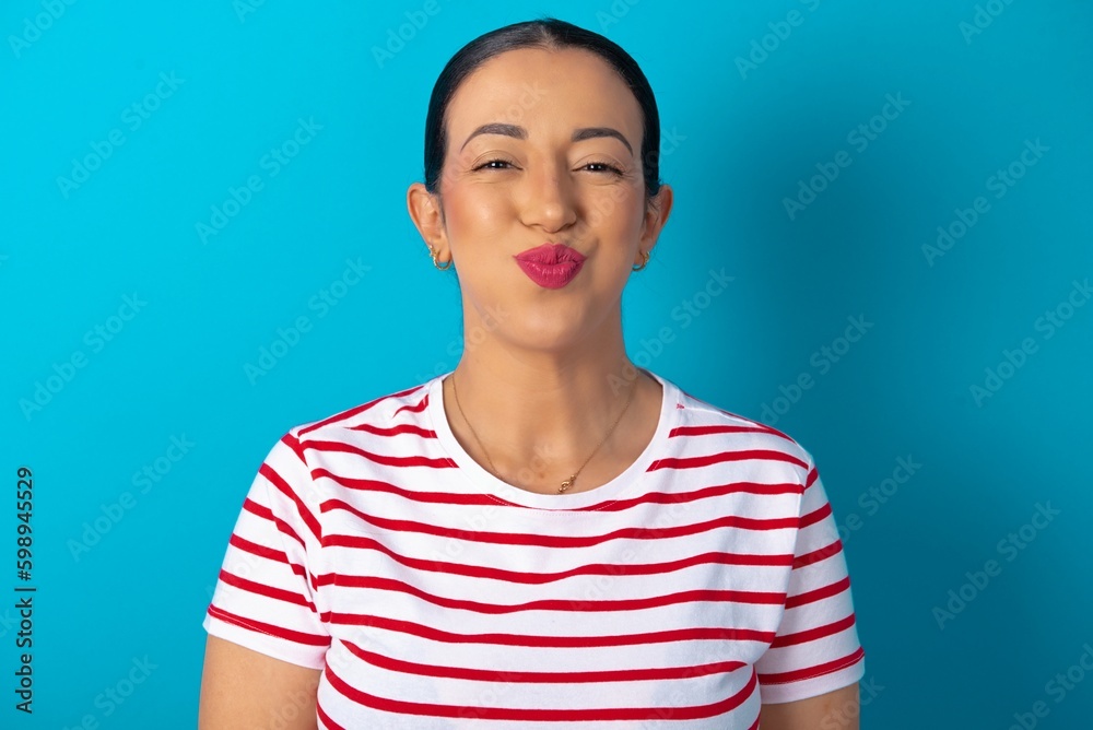beautiful woman wearing striped T-shirt over blue studio background puffing cheeks with funny face. Mouth inflated with air, crazy expression. beautiful woman wearing striped T-shirt over blue studio background puffing cheeks with funny face. Mouth inflated with air, crazy expression.