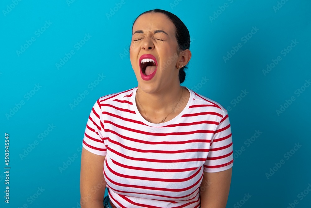 Stressful beautiful woman wearing striped T-shirt over blue studio background screams in panic, closes eyes in terror, keeps hands on head, finds out terrified news, can't believe it. Stressful beautiful woman wearing striped T-shirt over blue studio background screams in panic, closes eyes in terror, keeps hands on head, finds out terrified news, can't believe it.