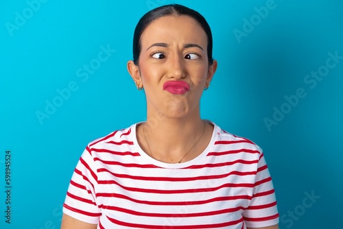 beautiful woman wearing striped T-shirt over blue studio background crosses eyes, puts lips, makes grimace with awkward expression has fun alone, plays fool.