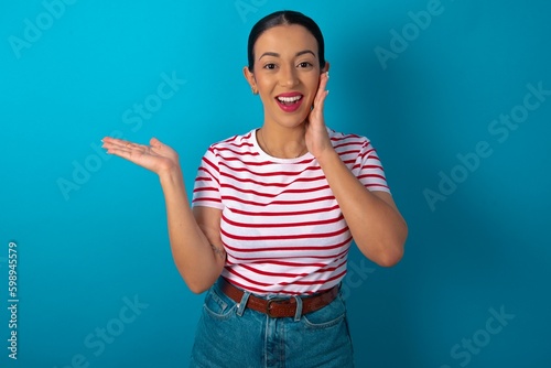 Positive glad beautiful woman wearing striped T-shirt over blue studio background says: wow how exciting it is, indicates something. One hand on his head and pointing with other hand.