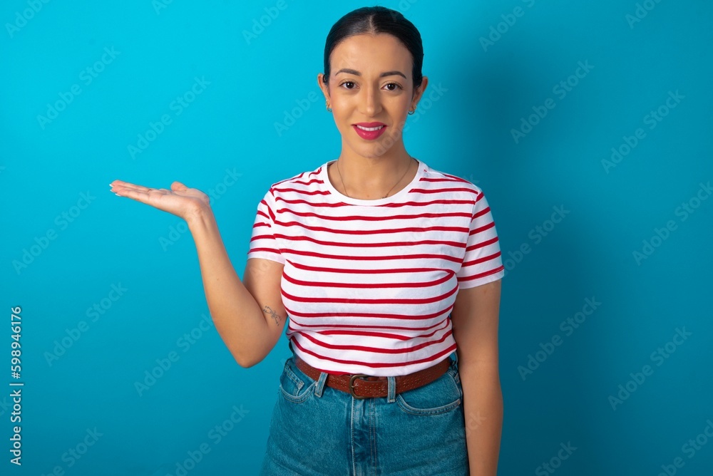 Positive glad beautiful woman wearing striped T-shirt over blue studio says: wow how exciting it is, has amazed expression, shows something on blank space with open hand. Advertisement concept. Positive glad beautiful woman wearing striped T-shirt over blue studio says: wow how exciting it is, has amazed expression, shows something on blank space with open hand. Advertisement concept.
