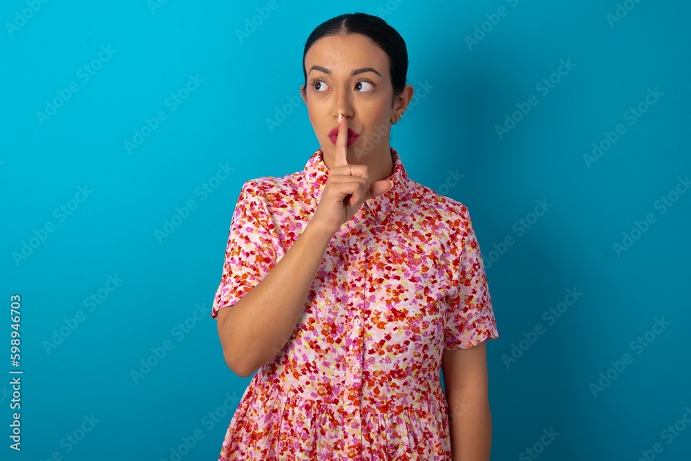 beautiful woman wearing floral dress over blue studio background makes silence gesture, keeps index finger to lips makes hush sign. Asks not to share secret. beautiful woman wearing floral dress over blue studio background makes silence gesture, keeps index finger to lips makes hush sign. Asks not to share secret.