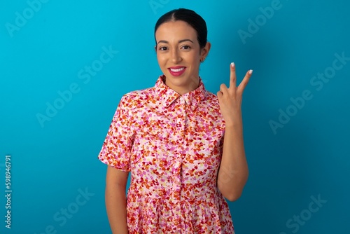 beautiful woman wearing floral dress over blue studio background smiling and looking friendly, showing number two or second with hand forward, counting down
