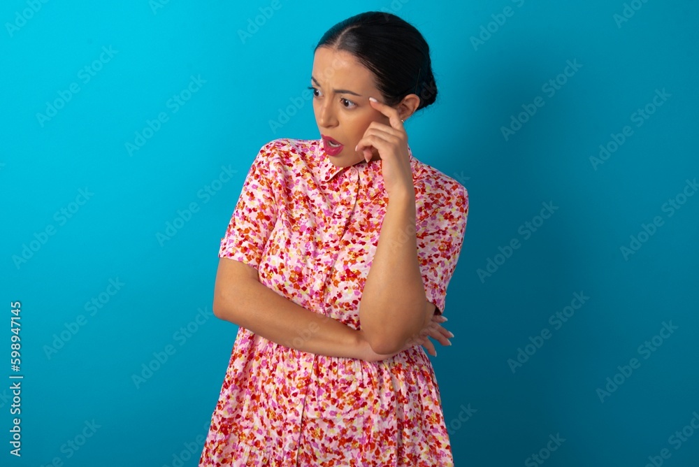 Astonished beautiful woman wearing floral dress over blue studio background looks aside surprisingly with opened mouth. Astonished beautiful woman wearing floral dress over blue studio background looks aside surprisingly with opened mouth.