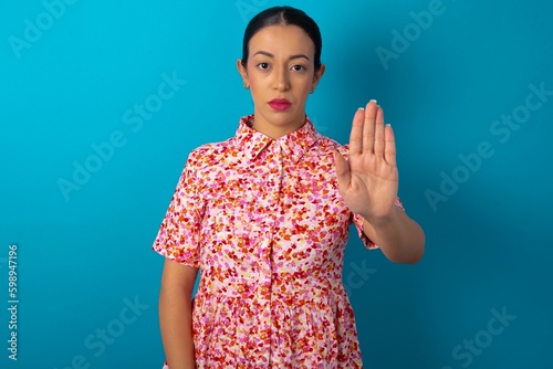 beautiful woman wearing floral dress over blue studio background shows stop sign prohibition symbol keeps palm forward to camera with strict expression