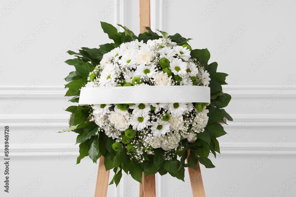 Funeral wreath of flowers with ribbon on wooden stand near white wall