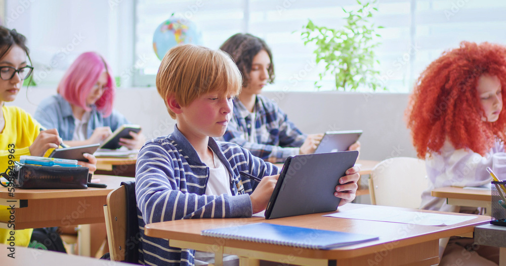 Small Caucasian schoolboy sitting at desk in classroom among kids at lesson and using tablet device while learning. Smart boy texting on screen of computer and looking at blackboard at school. Small Caucasian schoolboy sitting at desk in classroom among kids at lesson and using tablet device while learning. Smart boy texting on screen of computer and looking at blackboard at school.