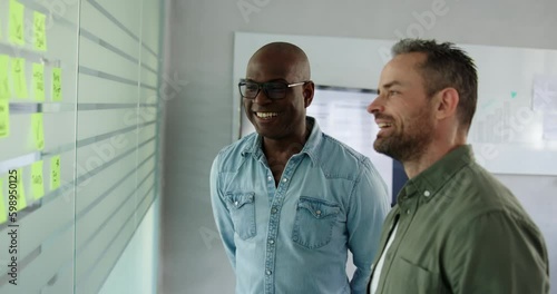 Two diverse businessmen laughing during a brainstorming session together with adhesive notes on a glass wall of an office