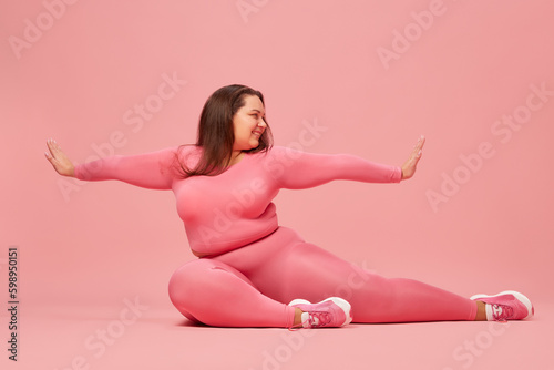 Well-being and self-love. Young overweight woman training in pink sportswear, doing stretching against pink studio background. Concept of sport, body-positivity, weight loss, body and health care