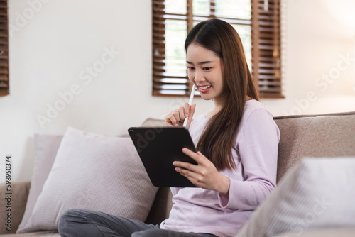 Beautiful attractive young Asian woman sitting on gray sofa and using digital tablet at living room in the morning