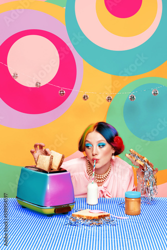 Beautiful young girl with bright makeup and colorful hair drinking milk, eating toast with confetti against abstract multicolored background. Party and celebration. Food pop art. Concept of creativity