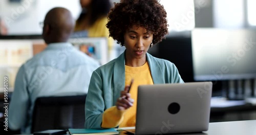 Young businesswoman smiling during a video call on a laptop while sitting at her desk in an office with colleagues in the background
