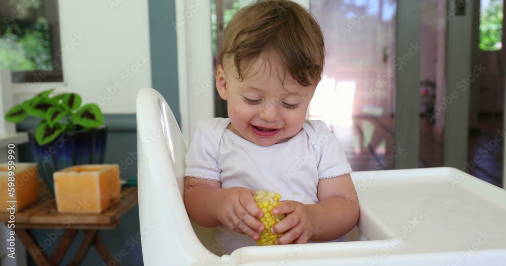 Happy adorable baby on highchair holding healthy corn Happy adorable baby on highchair holding healthy corn