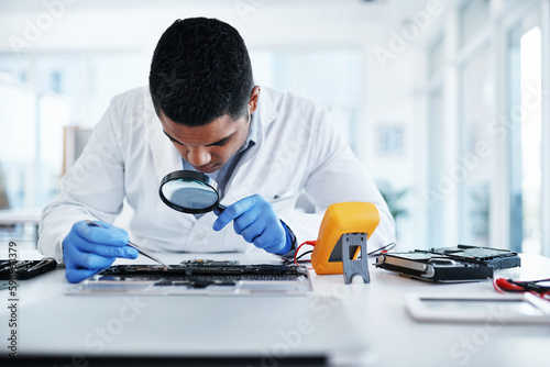 The surgeon of IT systems. a young man using tweezers and a magnifying glass while repairing computer hardware in a laboratory.