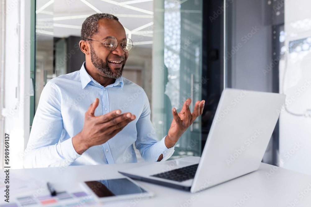 Mature african american boss working inside office with laptop remotely, businessman in shirt on online meeting with colleagues, man talking on video call using laptop while sitting in shirt. Mature african american boss working inside office with laptop remotely, businessman in shirt on online meeting with colleagues, man talking on video call using laptop while sitting in shirt.