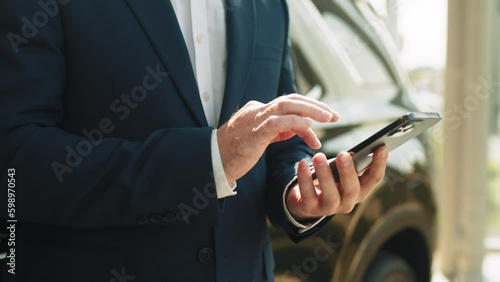 Middle age customer standing near new car in auto showroom. Making his choice. Close up of unrecognizable business man wearing blue formal suit standing next to car in showroom using his smartphone