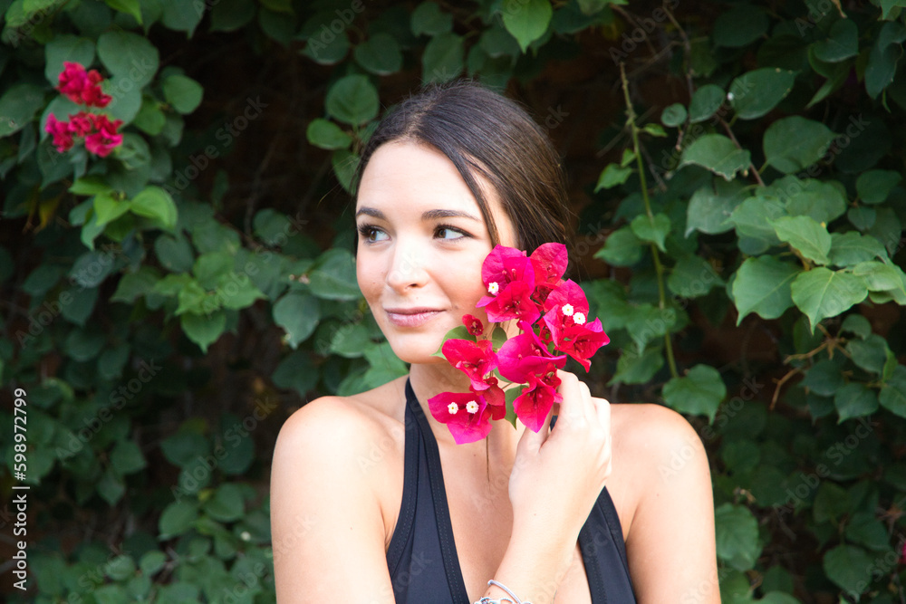 Young beautiful brunette typical Spanish woman dressed in black suit and bouquet of pink flowers near her face. The woman is in the park surrounded by greenery. The woman is happy. Young beautiful brunette typical Spanish woman dressed in black suit and bouquet of pink flowers near her face. The woman is in the park surrounded by greenery. The woman is happy.