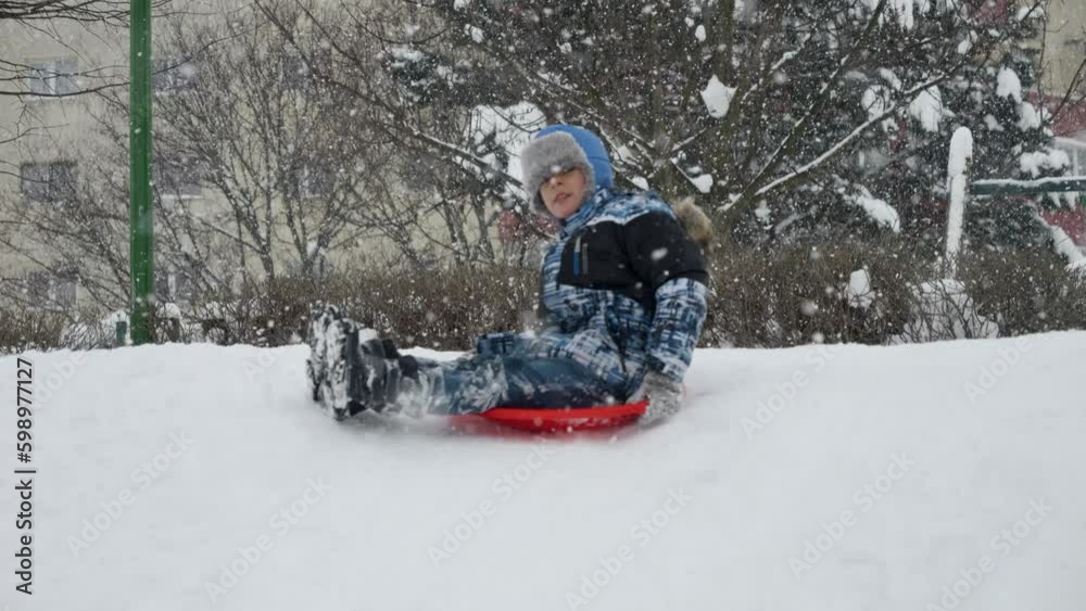 Slow motion of cheerful smiling boy riding down the snowy hill on red plastic sleds during snowfall Slow motion of cheerful smiling boy riding down the snowy hill on red plastic sleds during snowfall