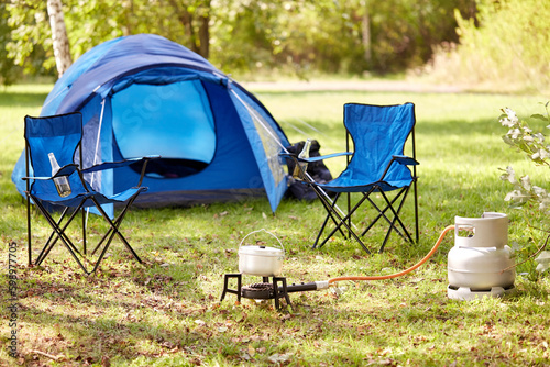 food cooking, tourism and travel concept - close up of camping pot on tourist gas burner, blue tent and two folding chairs at camp