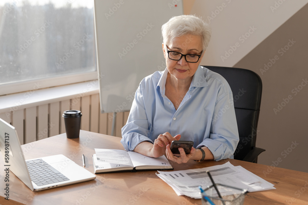 Confident stylish european middle aged senior woman using smartphone at workplace. Stylish older mature 60s gray haired lady businesswoman with cell phone in office. Boss leader using internet apps Confident stylish european middle aged senior woman using smartphone at workplace. Stylish older mature 60s gray haired lady businesswoman with cell phone in office. Boss leader using internet apps